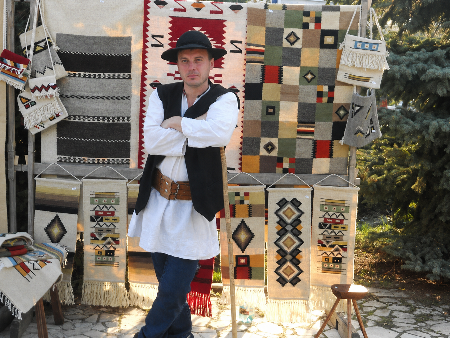 Iulian Mihalachi wearing a white shirt, black vest, blue jeans, and a black hat stands in front of a display of colorful traditional woven rugs and wall hangings.