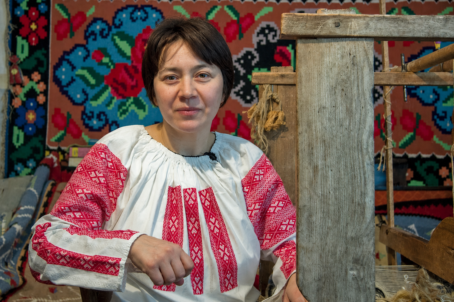 Maria Mihalachi wearing a traditional white Romanian embroidered blouse with red patterns, seated in front of a colorful woven rug and a wooden loom.