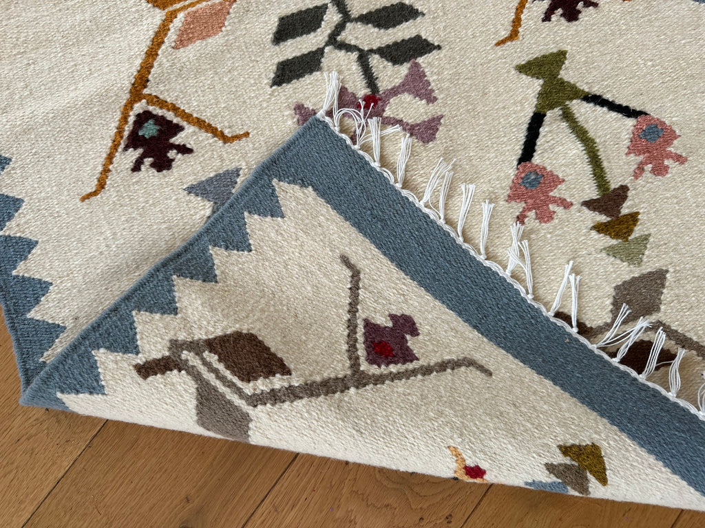 A close-up of a patterned area rug corner with white fringe, shown on a light wood floor. The rug has an off-white base with geometric borders in muted blue-gray and abstract motifs in brown, pink, olive green, and black.