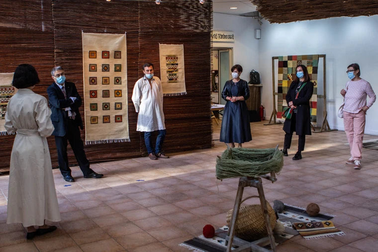 A group of people standing in a gallery for an exhibition with woven wall hangings and textile displays. In the foreground, a small wooden stand holds a knotted green bundle with nearby craft materials on the floor.